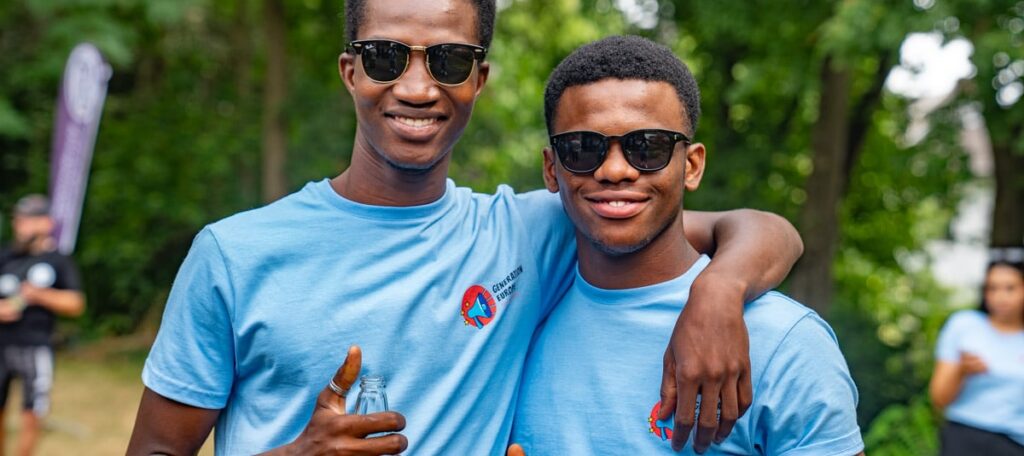 Two young people in light blue Generation Europe T-shirts and sunglasses stand arm in arm in front of trees.