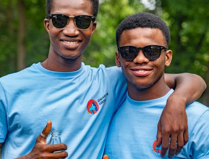 Two young people in light blue Generation Europe T-shirts and sunglasses stand arm in arm in front of trees.
