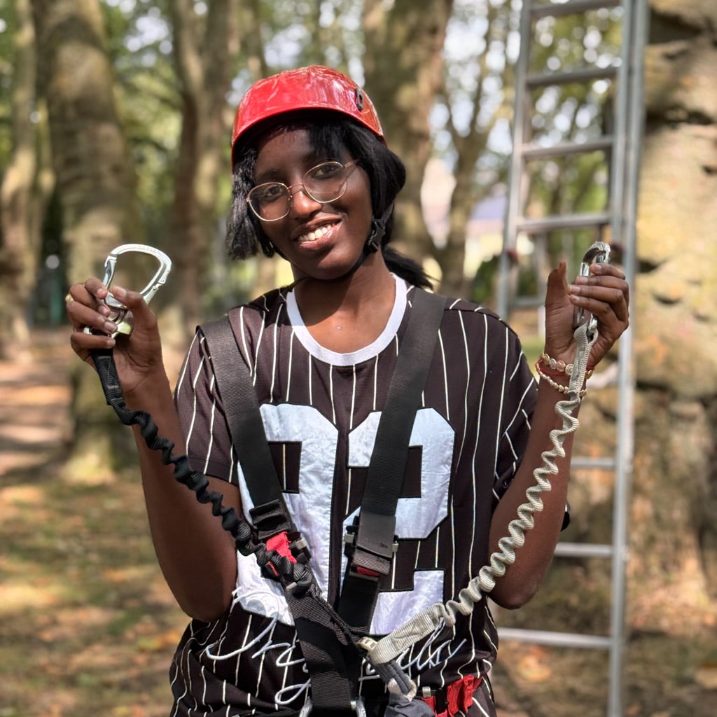 A young person wearing a red climbing helmet and safety harness holds two carabiners with safety lines on a high ropes course.