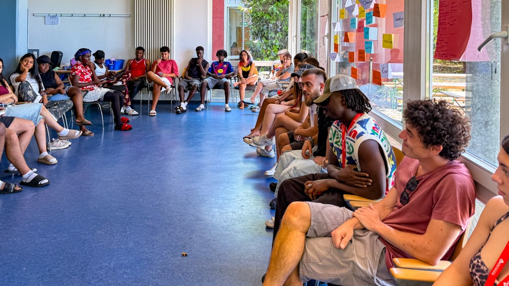 Around 20 young people sit in a circle with papers and notepads in a bright seminar room.